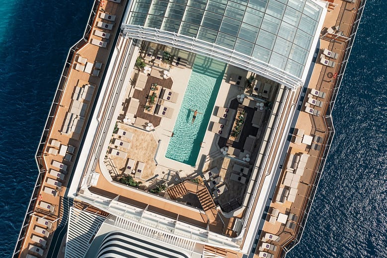 Luxury cruise ship deck viewed from above, showing a pool, lounge seating, and open ocean surrounding the vessel.