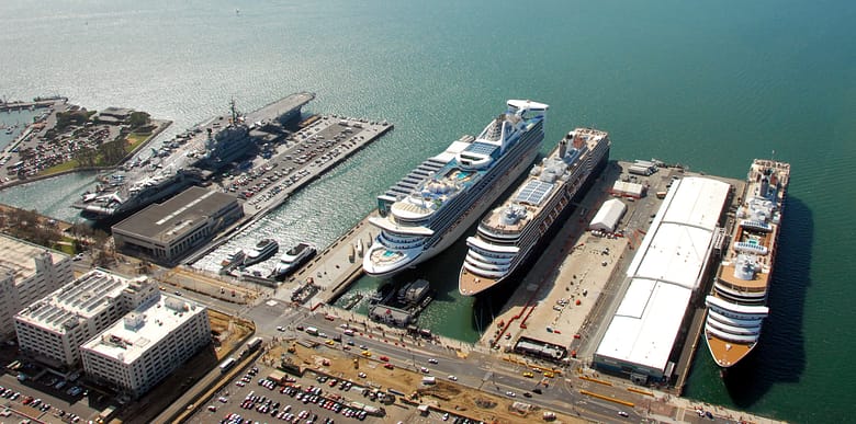 Aerial view of three large cruise ships docked at the San Diego cruise terminal, with the harbor, open water, and shoreline in the background.