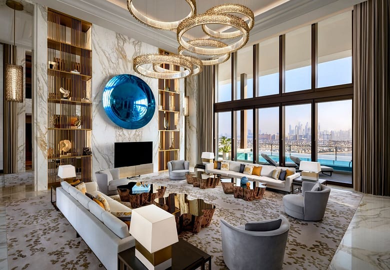 Luxury hotel suite with soaring ceilings, crystal ring chandeliers, marble walls, designer seating, and floor-to-ceiling windows overlooking a private terrace pool and city skyline.