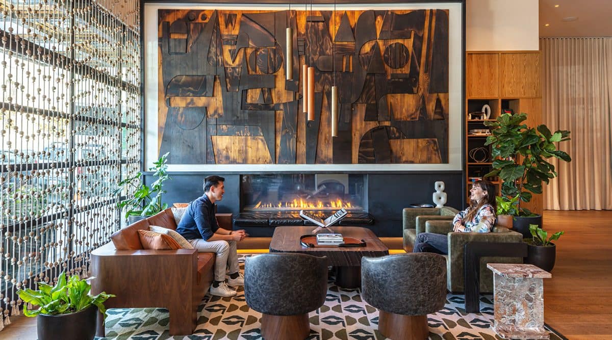 Luxury hotel lounge with contemporary art, fireplace, wood furnishings, and two guests seated in conversation surrounded by warm modern design.