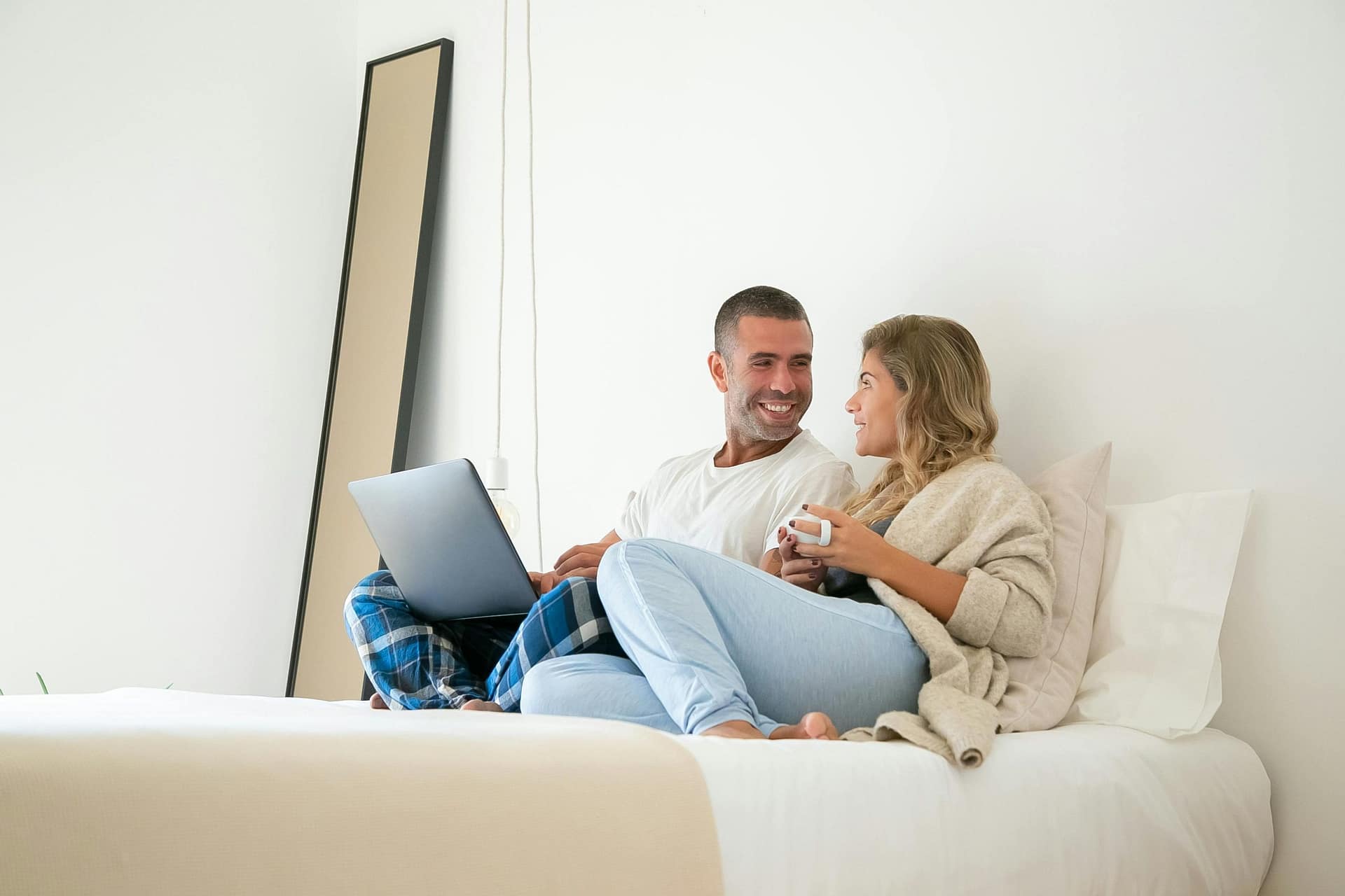 Couple sitting on a bed with a laptop, appearing to browse travel or hotel options together from a bedroom.