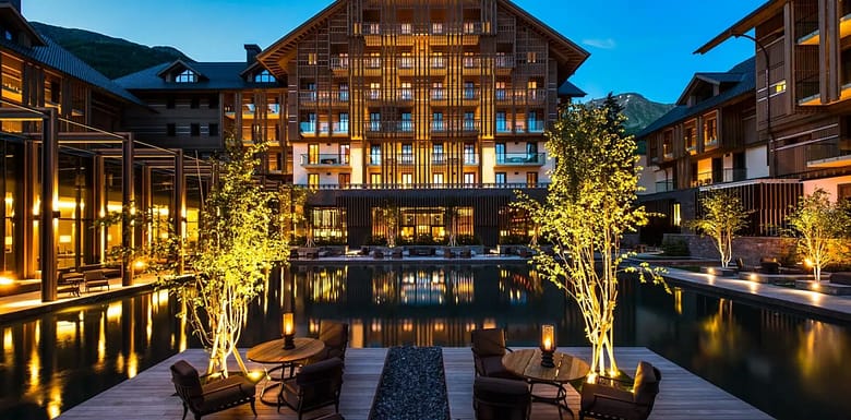 Illuminated luxury hotel exterior at dusk with warm lights reflecting across a still pool, framed by modern terraces, lounge seating, and mountain peaks in the background.