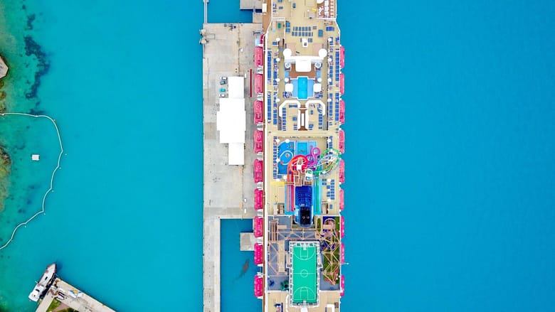 Aerial view of a large modern cruise ship docked at a port terminal with turquoise water and the ship’s upper decks visible from above.
