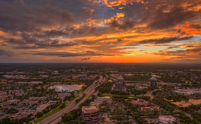 Aerial view of West Palm Beach, Florida at sunset with highways, office buildings, lakes, and residential areas under dramatic cloud cover.