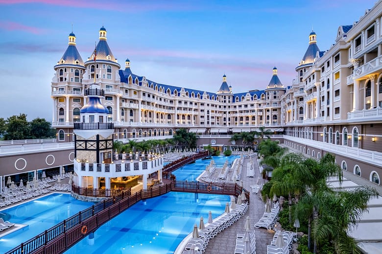 Luxury resort hotel courtyard with ornate architecture, blue-roofed towers, and a large swimming pool complex surrounded by lounge chairs and palm trees at dusk.