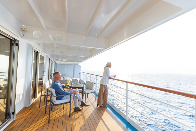Luxury cruise ship suite veranda with a couple relaxing by the ocean, seated and standing on a spacious private balcony with outdoor dining table and sea views.