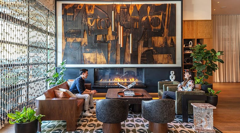 Luxury hotel lounge with contemporary art, fireplace, wood furnishings, and two guests seated in conversation surrounded by warm modern design.