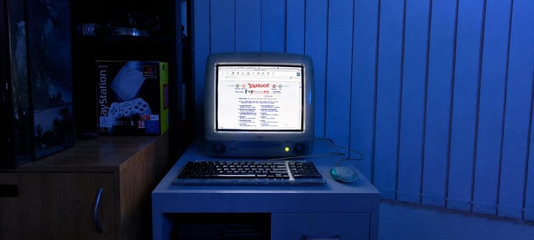 A late 1990s iMac displaying Yahoo on screen with a PlayStation box visible in the background, representing the early internet era when Americas Great Resorts published its 1998 warning about OTA dependency in luxury hospitality.