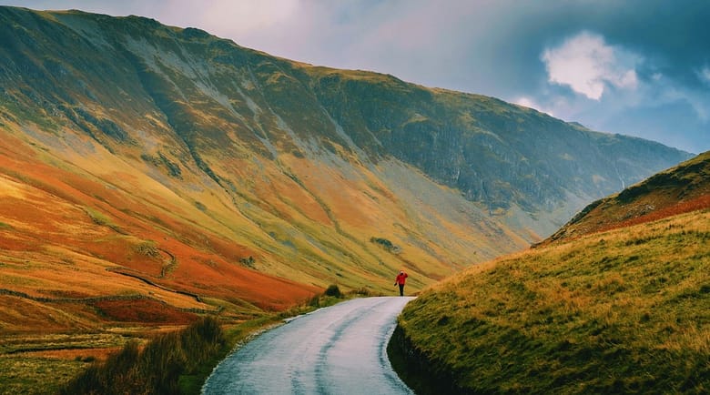 A lone traveler walks a narrow road toward a towering mountain range, illustrating the gap between confidence in direction and understanding of the scale of what lies ahead.