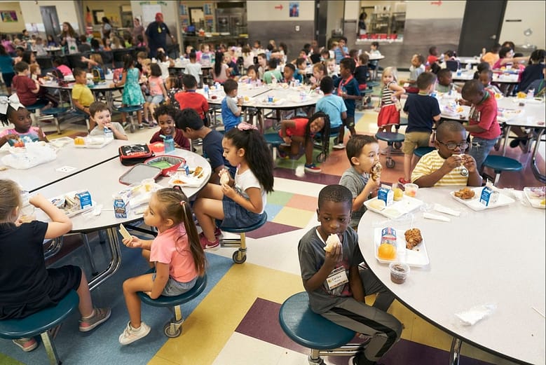 A crowded elementary school cafeteria filled with children eating lunch, illustrating the competitive environment hotels navigate as AI systems reshape who controls the room where travel decisions are made.
