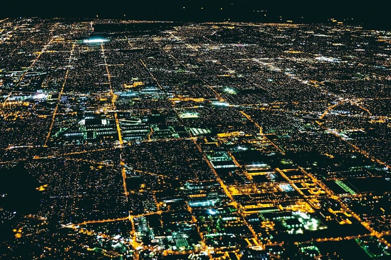 Aerial night view of a brightly lit city grid, representing the complex information environment AI systems use to discover and recommend hotels.