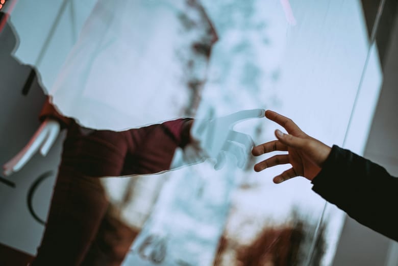 Hand reaching toward a reflected image through glass, representing mediated digital interaction in AI-driven hotel discovery.