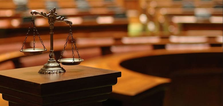Scales of justice on a courtroom podium with empty wooden benches in the background.