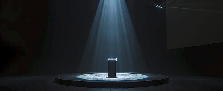 An empty stage with a single spotlight illuminating a podium in a dark room — the infrastructure for an audience that was never acquired.